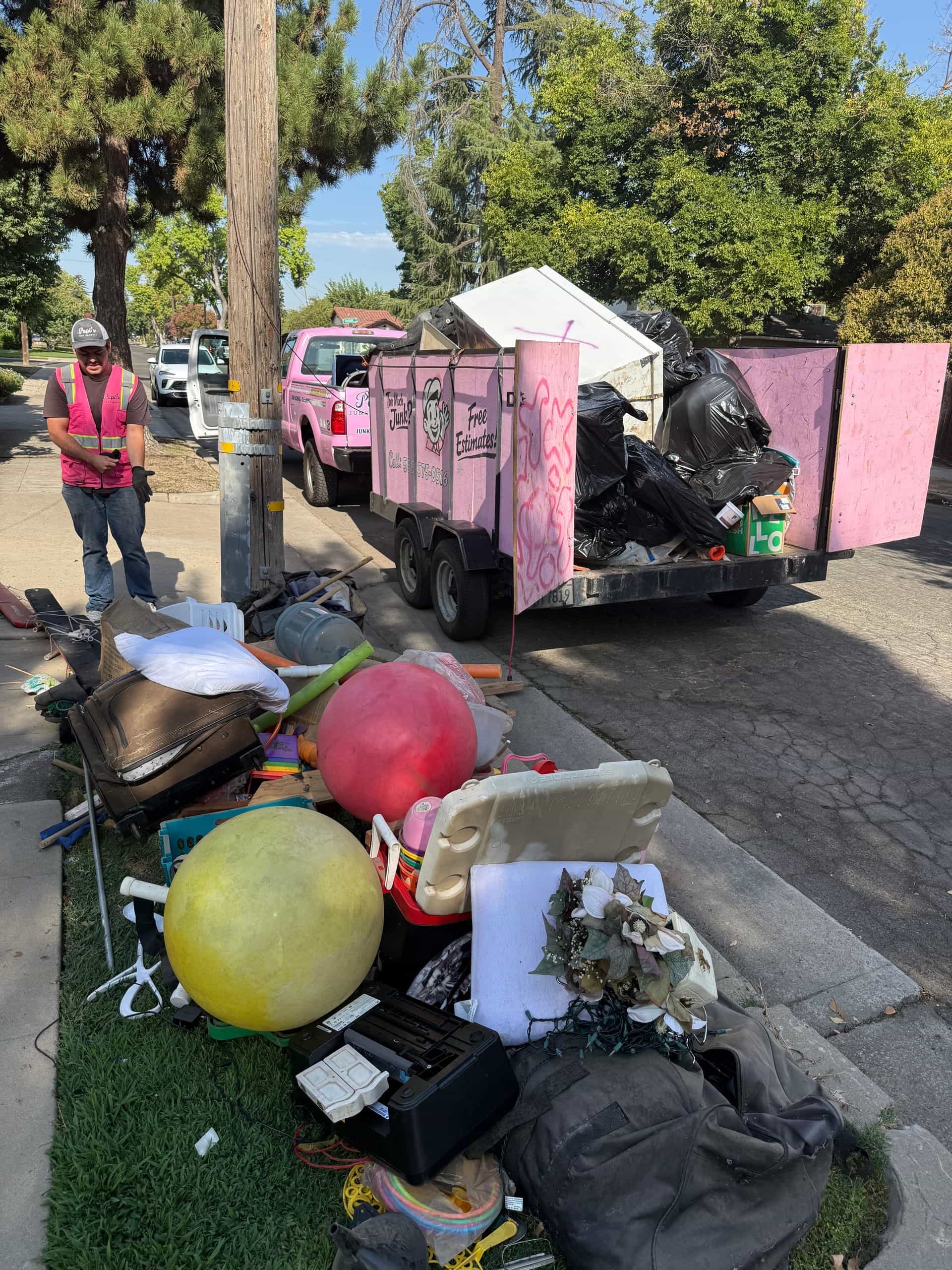 Curbside pile of household junk beside Papi's pink trailer and truck in Fresno, crew member in a pink vest