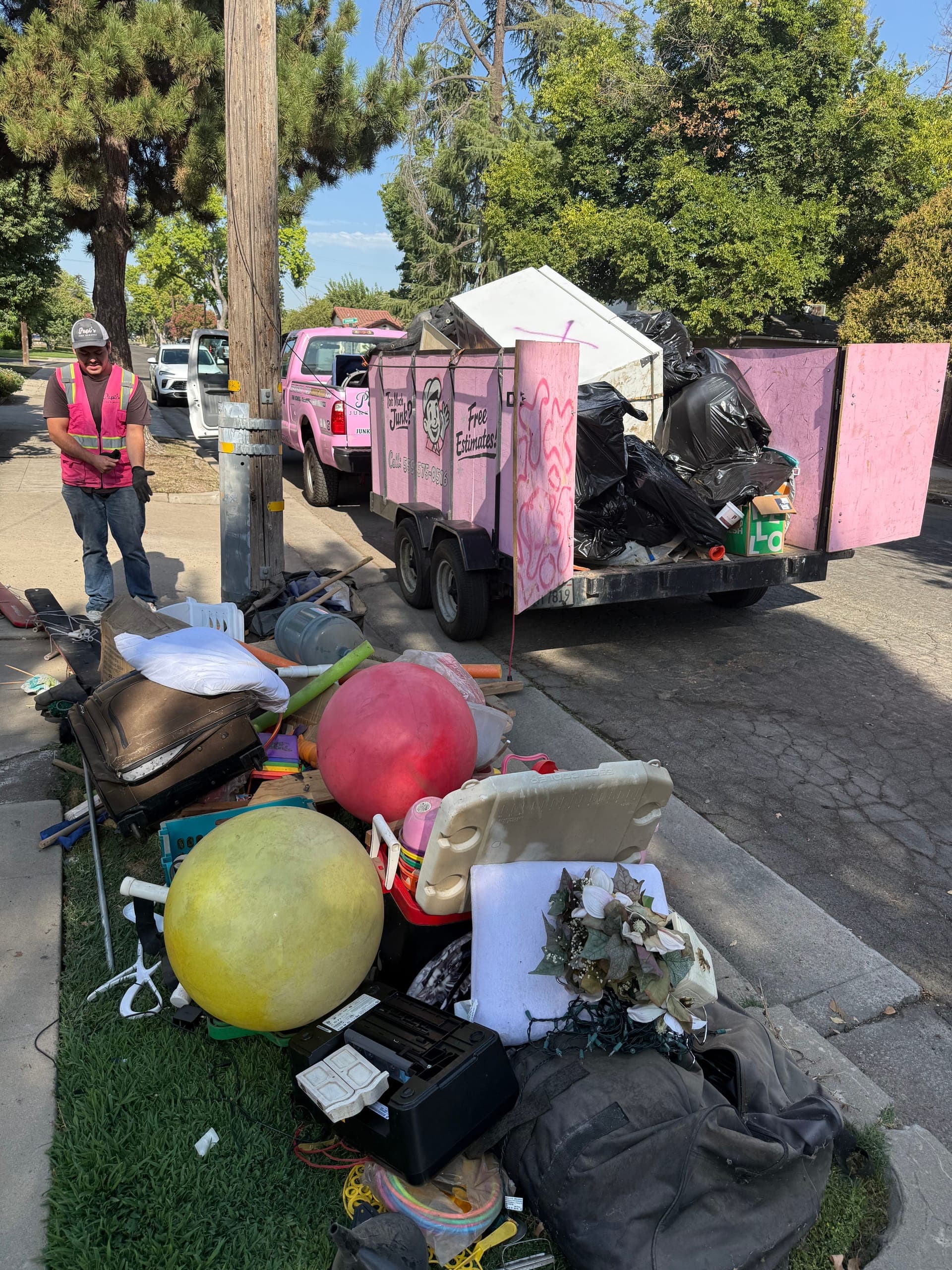 Curbside pile of household junk beside Papi's pink trailer and truck in Fresno, crew member in a pink vest
