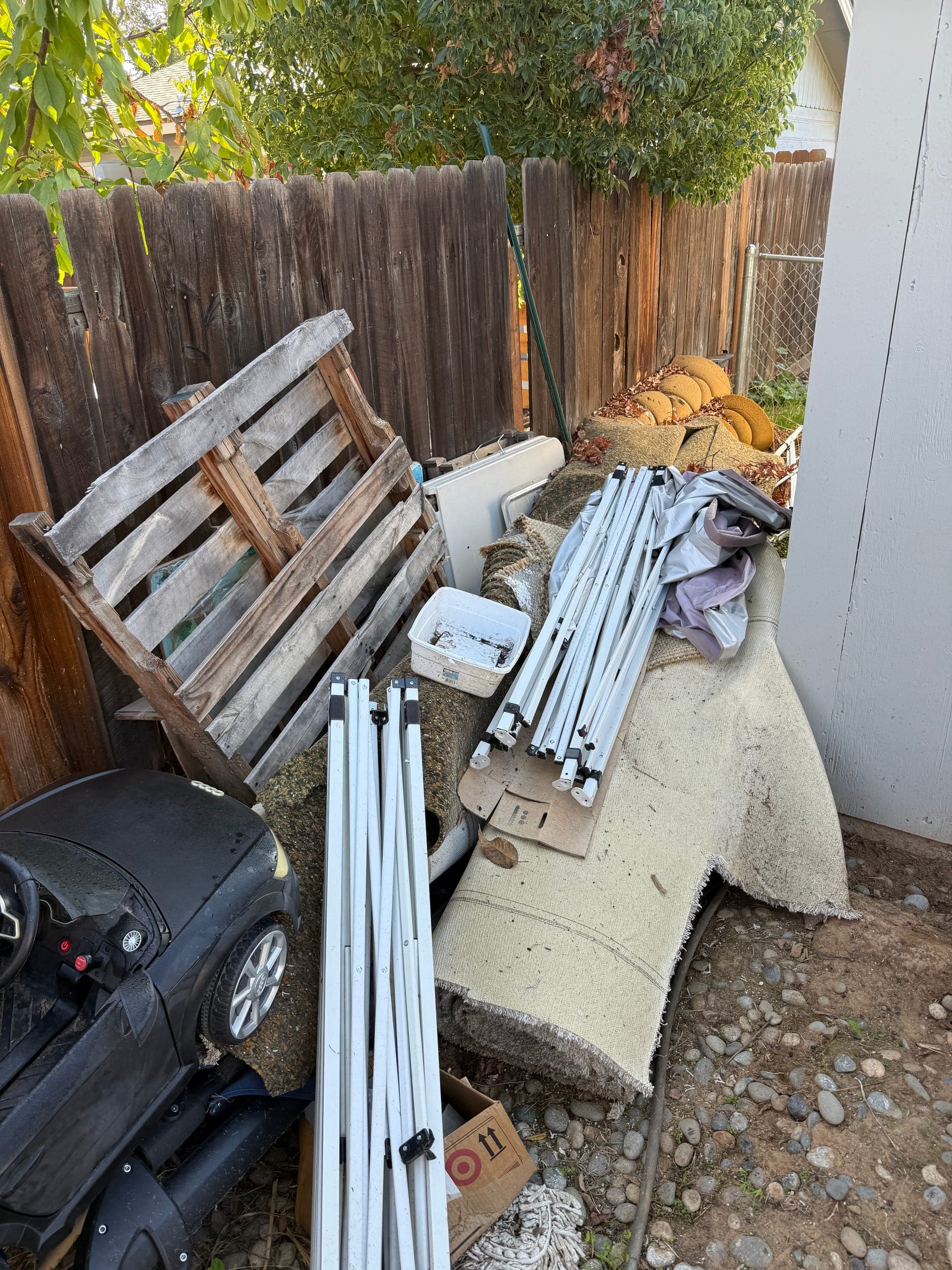 Side yard crowded with pallets, carpet, canopy poles and a kids' ride-on car before cleanup